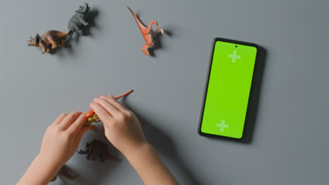 Overhead-Shot-Of-Boy-Playing-With-Toy-Dinosaurs-Next-To-Green-Screen-Mobile-Phone